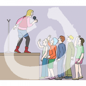 Eine Person mit Mikrofon in der Hand steht auf einer Bühne. Sie guckt zu fünf Menschen runter.