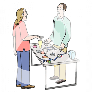 Eine Person steht hinter einem Tisch mit verschiedenen Gegenständen. Sie zeigt auf eine Uhr. Davor steht eine weitere Person und streckt die Handfläche nach oben.