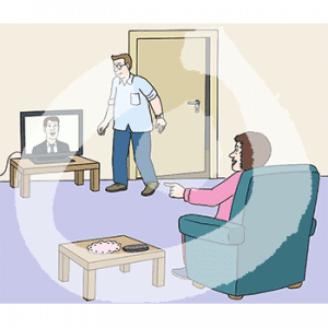 Eine Person sitzt auf einem Sessel. Sie zeigt auf den Bildschirm eines Fernsehers mit einem Mann darin. Daneben steht eine Person.