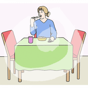 Eine Frau sitzt an einem Tisch, sie führt einen Löffel zum Mund. Vor ihr steht ein Becher und eine Schüssel mit Essen.