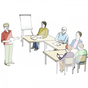 Fünf Personen sitzen um einen Tisch herum. Vor jeder liegt ein Blatt und ein Stift. Eine Person steht davor. Im Hintergrund ist ein Flipchart.