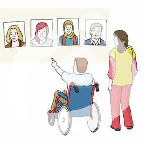 Eine Person im Rollstuhl zeigt auf eins von vier Portraits an einer Wand. Daneben steht eine weitere Person.