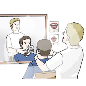 Eine Person sitzt vor einem Spiegel. Eine andere Person mit Handschuhen zieht von hinten den Mund auseinander. Neben dem Spiegel hängen Bilder von Mundpositionen und den jeweiligen Buchstaben.