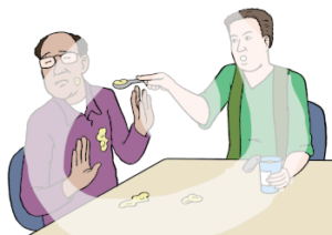 Zwei Personen sitzen an einem Tisch. Eine Person hält in einer Hand einen Becher, in der anderen Hand hat sie einen Löffel mit Essen. Sie hält den Löffel vor den Mund der anderen Person. Diese Person hebt abwehrend die Hände. Auf ihrem Oberteil und auf dem Tisch ist Essen.