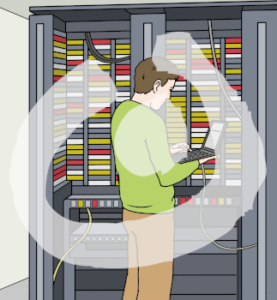 Eine Person steht vor einer schrankartigen Fläche. In dieser Fläche sind leuchtende Felder und Kabel. Die Person hält einen Laptop in der Hand.